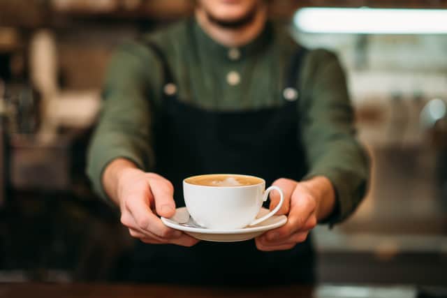 Waiter in black apron stretches a cup of coffee with cream in hands