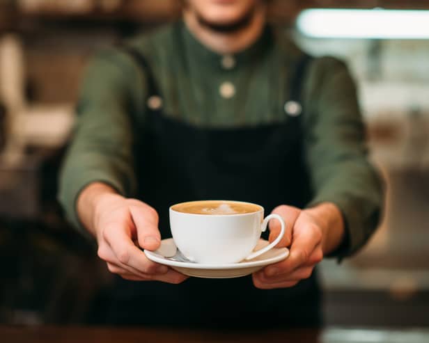 Waiter in black apron stretches a cup of coffee with cream in hands